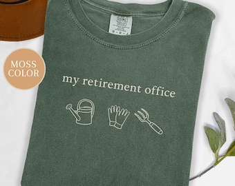 May include: A moss-colored t-shirt with the text "my retirement office" in white. Below the text are white line drawings of a watering can, gardening gloves, and a garden fork. A brown hat is visible in the background.