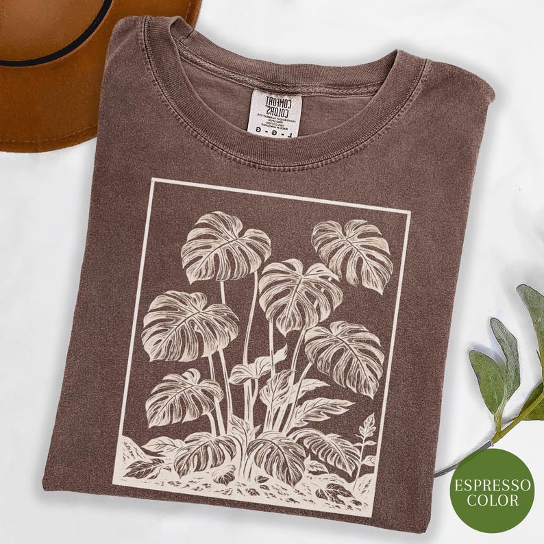 May include: Espresso-colored t-shirt featuring a white graphic of monstera leaves within a rectangular frame. The shirt has a crew neck and short sleeves. The tag reads "TROHMOJ 280J0J". The shirt is a neutral color and suitable for casual wear.