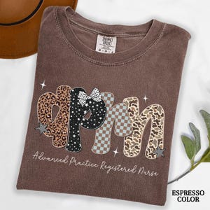 May include: A brown t-shirt with the letters "APRN" in a decorative font. The letters feature leopard print, stars, a bow, and a checkered pattern. The text "Advanced Practice Registered Nurse" is below the letters.
