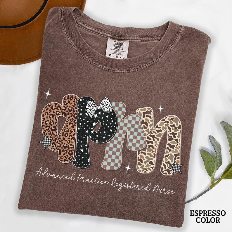 May include: A brown t-shirt with the letters "APRN" in a decorative font. The letters feature leopard print, stars, a bow, and a checkered pattern. The text "Advanced Practice Registered Nurse" is below the letters.