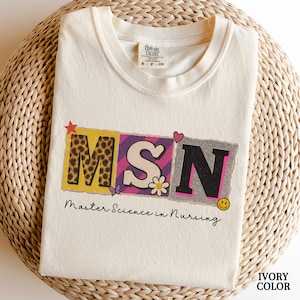 May include: Ivory-colored t-shirt with the letters "MSN" in a colorful, collage-style design. The "M" has a leopard print, the "S" has a flower, and the "N" has a smiley face. The text "Master Science in Nursing" is below.
