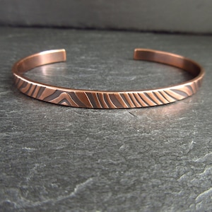 May include: Copper cuff bracelet with an etched, textured design. The bracelet has a warm, reddish-brown colour and a simple, open design. The etched pattern features a series of curved lines, creating a unique visual texture. This piece of jewellery is a stylish accessory.