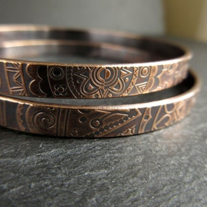 Two bronze metal bangles sit on top of each other on a piece of grey slate. The metal has a detailed doodle style pattern imprinted into the surface.