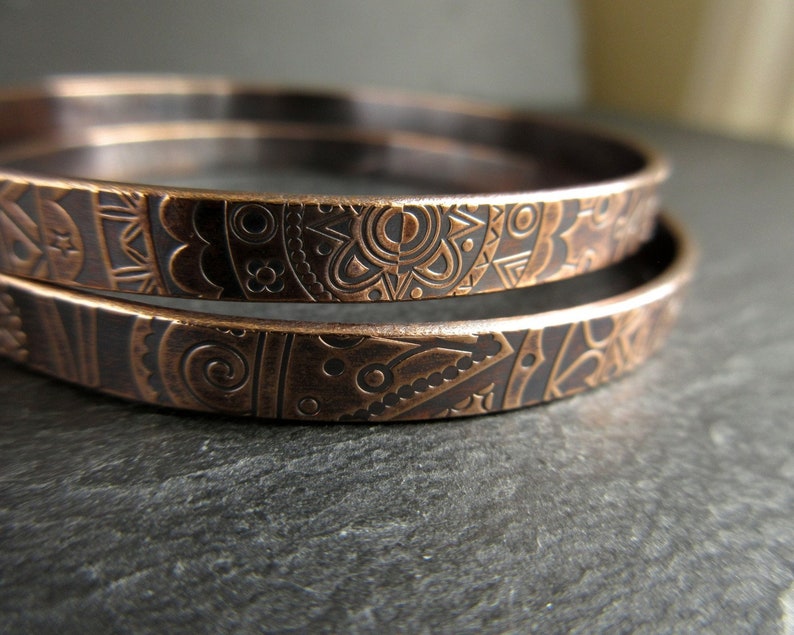 Two bronze metal bangles sit on top of each other on a piece of grey slate. The metal has a detailed doodle style pattern imprinted into the surface.