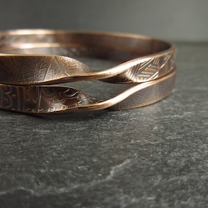 May include: Two copper bracelets with intricate etched designs. The bracelets are stacked on top of each other and have a wavy, organic shape.
