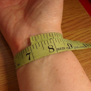 close up of inside of wrist with green tape measure wrapped around wrist to demonstrate how to measure for a cuff bangle.