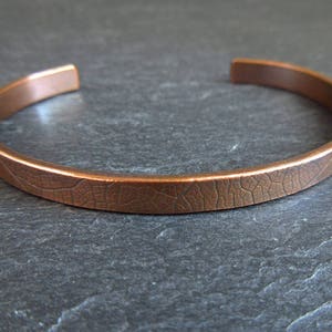 May include: A copper bracelet with a textured, leaf-like pattern. The bracelet is a simple, minimalist design.