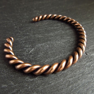 A close up of a bronze cuff bangle  sitting at an angle on a piece of slate. The bangle is made of two pieces of wire twisted together. It has an oxidised finished.