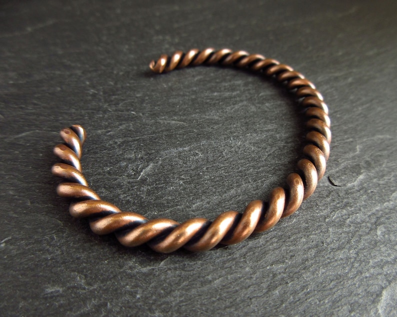 A close up of a bronze cuff bangle  sitting at an angle on a piece of slate. The bangle is made of two pieces of wire twisted together. It has an oxidised finished.