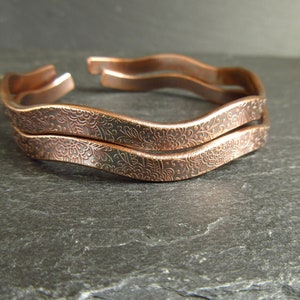 May include: Two copper bracelets with a wavy design and an intricate floral pattern.