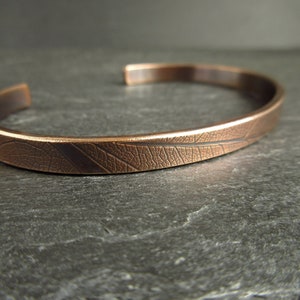 A close up of a bronze cuff bangle showing the leaf vein texture. The colour is a warm brown and the cuff sits on a piece of grey slate.