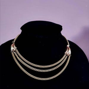 May include: Gold-toned necklace with three layered chain strands. The necklace features a decorative element at the top with a curved design. The necklace is displayed on a black mannequin head against a purple background.