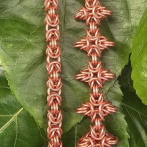 Puede incluir: Dos pendientes de cota de malla de color cobre. Un pendiente es una cadena simple, el otro presenta un diseño de cruz. Los pendientes se muestran sobre un fondo de hojas verdes. Los pendientes están hechos a mano.