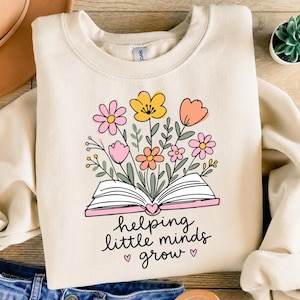 May include: A cream-colored crewneck sweatshirt featuring a graphic of an open book with colorful flowers in shades of pink, yellow, and orange emerging from the pages. Below the book, the text reads "helping little minds grow" with two small hearts.