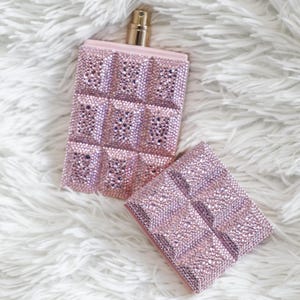 May include: A pink perfume bottle and matching case, both covered in sparkling rhinestones. The bottle has a gold-colored atomizer and a rectangular shape with a grid pattern of rhinestones. The case is also rectangular and has the same rhinestone grid design.