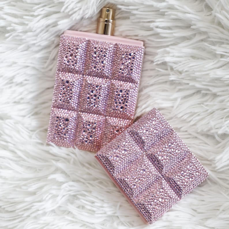 May include: A pink perfume bottle and matching case, both covered in sparkling rhinestones. The bottle has a gold-colored atomizer and a rectangular shape with a grid pattern of rhinestones. The case is also rectangular and has the same rhinestone grid design.