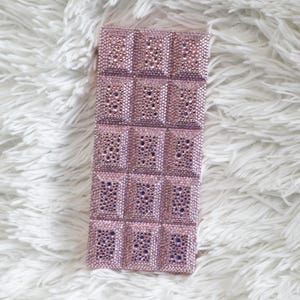 May include: A rectangular, pink, bedazzled phone case. The case is covered in small, sparkling rhinestones, arranged in a grid pattern. The case has a soft, plush texture.