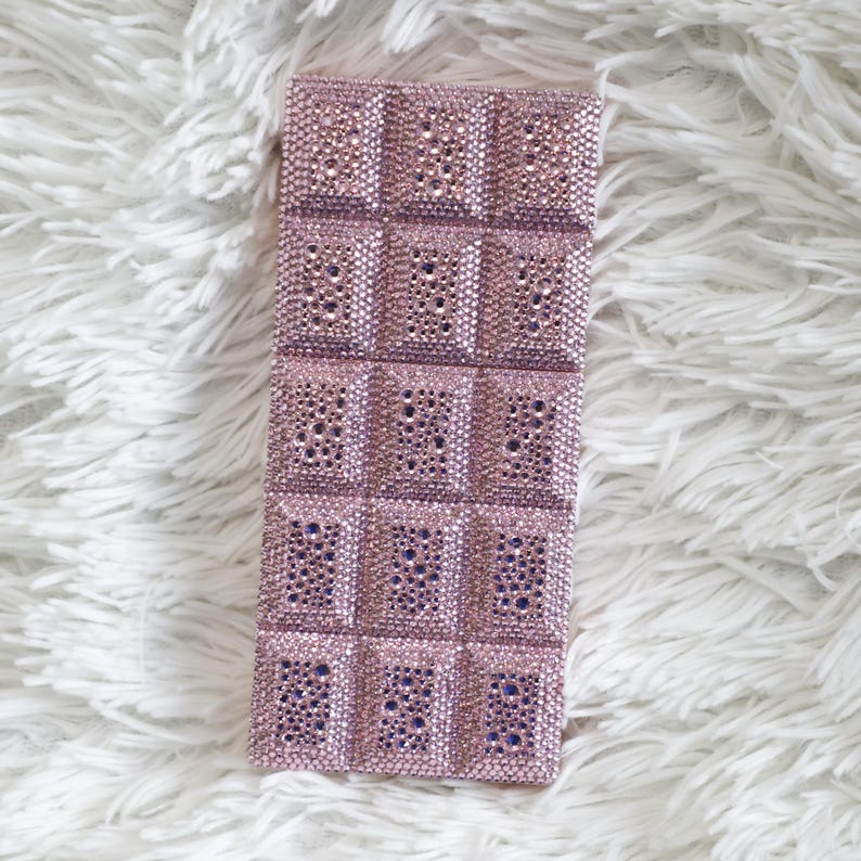 May include: A rectangular, pink, bedazzled phone case. The case is covered in small, sparkling rhinestones, arranged in a grid pattern. The case has a soft, plush texture.