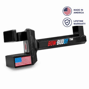 Op de afbeelding: Een zwarte Bow Buddy boogstandaard met een horizontale arm met de tekst BOW BUDDY TM in rood, wit en blauw. Het apparaat bevat een kleine Amerikaanse vlagsticker, een Made in America-pictogram en een schildpictogram voor een levenslange garantie.