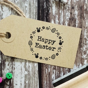 May include: A brown paper tag with a black stamp that says "Happy Easter" surrounded by a wreath of flowers, eggs, and bunny ears. A rainbow-coloured wooden stamp is in the background.