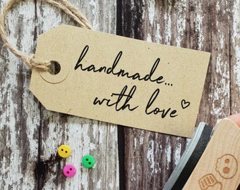 May include: A brown paper tag with the text "handmade... with love" written in black ink. A small heart symbol is next to the word "love". The tag is tied with twine and is sitting on a wooden surface. There are three colorful buttons on the surface.