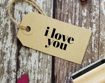 May include: A brown paper tag with a string attached, hanging on a wooden surface. The tag has the words "i love you" printed in black letters. There are two small pink hearts on the wood surface.