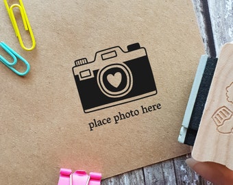 May include: A brown paper card with a black and white camera illustration and the text "place photo here". The camera has a heart in the lens. A wooden stamp with a cupcake and skull design is in the background.