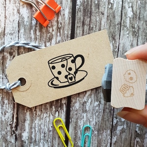 May include: A wooden stamp with a black ink pad. The stamp features a black and white illustration of a teacup with a teabag and a heart. The stamp is on a brown wooden surface.