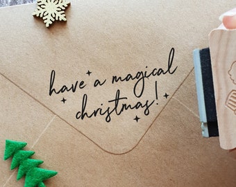 May include: A brown paper envelope with a wooden stamp that reads "have a magical christmas!" The stamp is being used to stamp the envelope. There are also small wooden decorations on the envelope, including a snowflake and a Christmas tree.