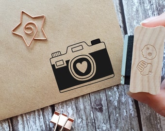 May include: A brown paper card with a black and white illustration of a camera with a heart in the lens. A gold star and a wooden stamp with a cupcake and skull design are also on the card.