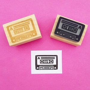 May include: Two wooden stamps featuring a vintage cassette tape design. The stamps are on a pink background. The cassette tape design is in black and white.