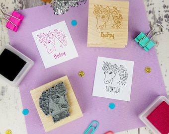 May include: A collection of unicorn-themed stamps and ink pads on a purple surface. The stamps are made of wood and rubber, featuring unicorn designs and names like "Betsy" and "Georgia." Pink and black ink pads are also present, along with colorful paper clips.