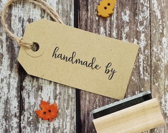 May include: A brown paper tag with a string attached and the text "handmade by" written in black cursive. The tag is sitting on a wooden surface with two small orange leaf-shaped buttons.