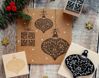 May include: Two wooden rubber stamps featuring intricate bird and foliage designs, one shaped like a Christmas ornament and one like a gift box. A brown card displays stamped impressions of these designs alongside white snowflake accents.
