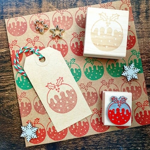 May include: A set of three wooden Christmas pudding stamps with holly leaves and red berries. The stamps are on a brown paper background with a red and green Christmas pudding pattern.