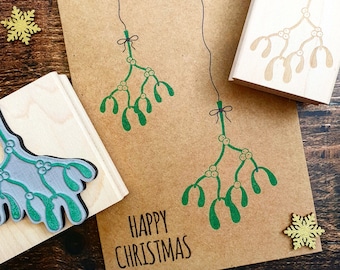 May include: A brown card with the text "Happy Christmas" printed on it. Two green mistletoe sprigs are printed on the card. The card is next to a wooden block with a green mistletoe sprig printed on it.