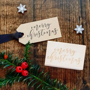 May include: Two wooden signs with the text "merry christmas" written in cursive. One sign is brown and the other is white. The signs are on a wooden surface with a green plant and red berries.