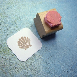 May include: A wooden stamp with a pink seashell design. The stamp is used to create a brown seashell imprint on a white card.