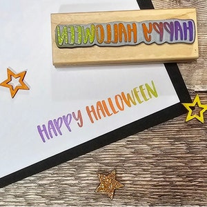 May include: A wooden stamp with the words "HAPPY HALLOWEEN" in a colorful gradient. The stamped image is in the same gradient. Decorative star cutouts in orange and gold glitter are scattered around the stamped image.
