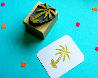 May include: A wooden stamp with a gold palm tree design. The stamp is on a blue surface and a white card with a gold palm tree imprint is next to it.