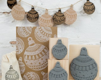 May include: A set of four gray rubber stamps featuring a Christmas ornament design. The stamps are mounted on wooden blocks and are arranged in a row. The stamps are surrounded by a brown paper bag, a brown card with a Christmas ornament design, and a string of Christmas ornaments.