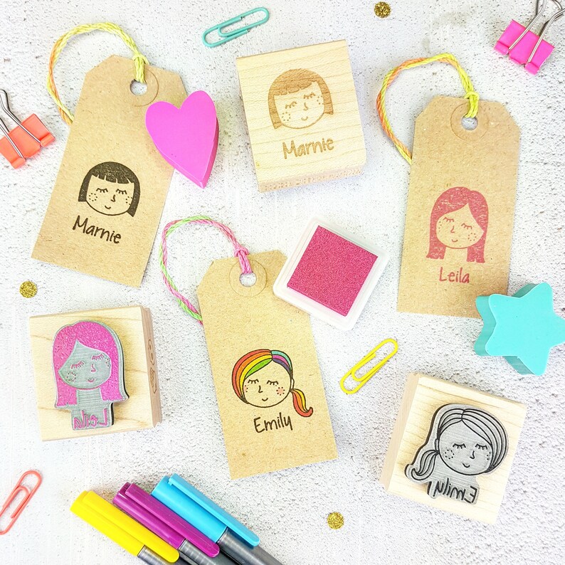 May include: A set of six wooden stamps featuring different girl faces with names, including Marnie, Leila, Emily, and Millie. The stamps are on a white background with colorful accents, including pink, blue, and yellow.