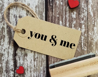 May include: A brown paper tag with the text "you & me" in black lettering. The tag is tied with twine and is hanging on a wooden surface. There are two red heart shapes on the wood.