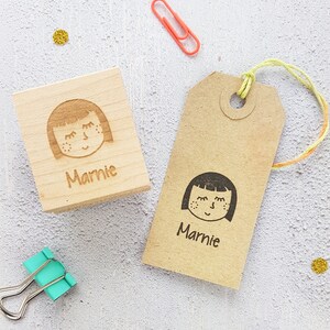 May include: Two wooden stamps with a girl's face and the name "Marnie" engraved on them. One stamp is on a wooden block and the other is on a brown paper tag.