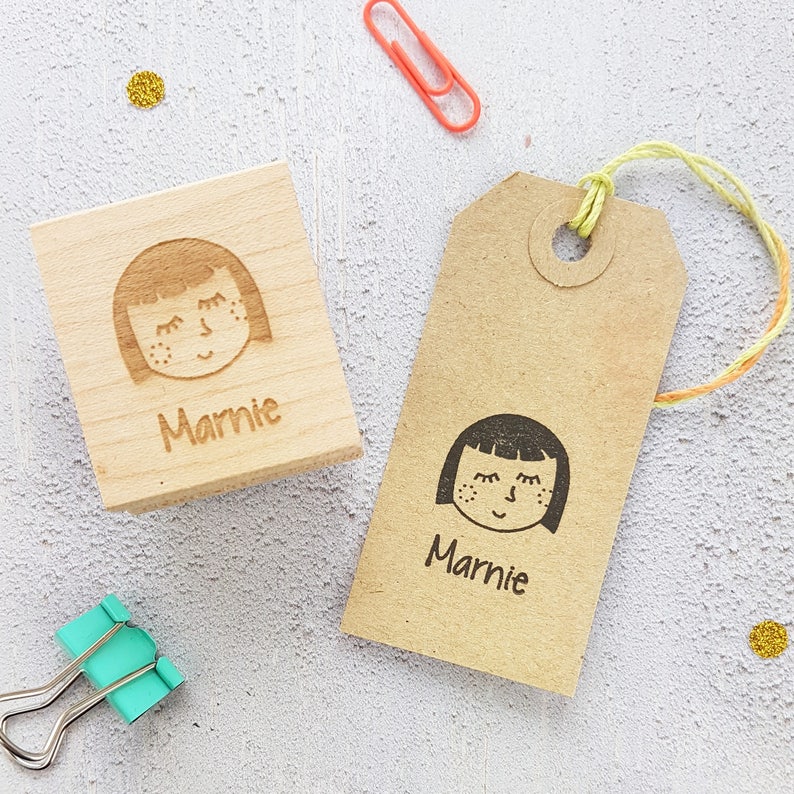 May include: Two wooden stamps with a girl's face and the name "Marnie" engraved on them. One stamp is on a wooden block and the other is on a brown paper tag.