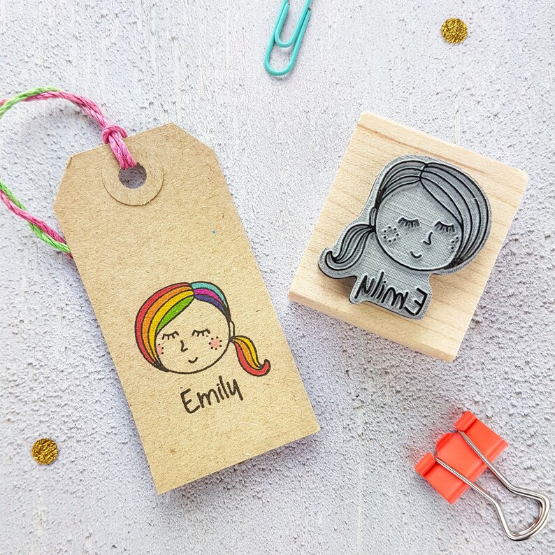 May include: A brown paper tag with a rainbow-haired girl illustration and the name "Emily" printed below. A wooden block stamp with a girl's face and the name "Emily" printed on it.