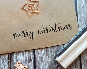 Könnte beinhalten: Ein beigefarbener Papierumschlag mit dem schwarzen Aufdruck merry christmas, einer kupferfarbenen Draht-Büroklammer in Sternform, einem Holzstempel und einer kupferfarbenen Metall-Klammer auf einer grauen Holzoberfläche.
