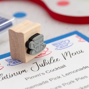 May include: A wooden stamp with a grey rubber design of a teacup with a Union Jack pattern. The stamp is on top of a blue and white card with the text "Platinum Jubilee Menu" and a list of menu items.