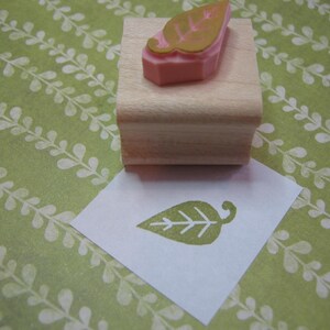 May include: A wooden block with a pink and gold leaf design rubber stamp. The stamp is used to create a leaf impression on a white piece of paper.