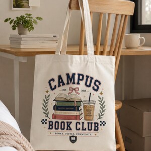 May include: A cream-colored canvas tote bag featuring a graphic print with the text CAMPUS BOOK CLUB. The design includes a stack of four books, a pink bow, a plastic cup of iced coffee, and green leafy branches, with the text BOOKS. COFFEE. COMMUNITY.